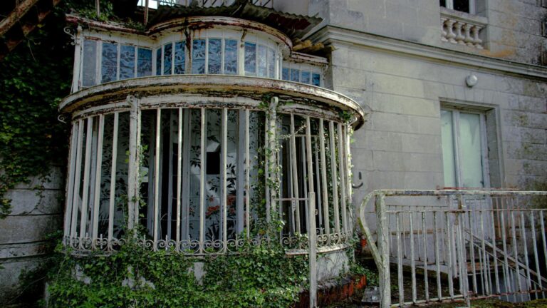 Dilapidated curved glass conservatory with ornate ironwork and climbing ivy on a historic stone building