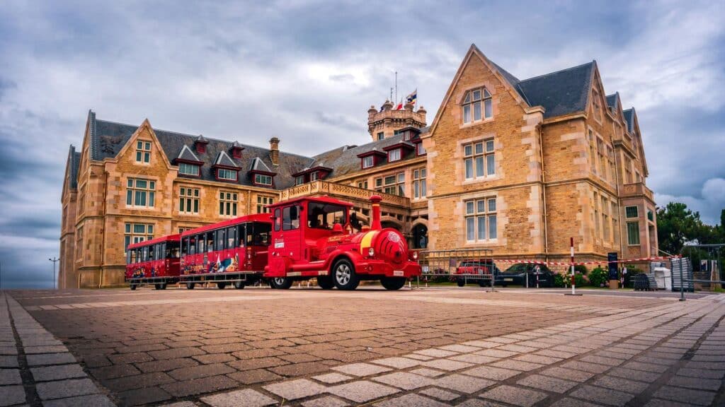 Red tourist train parked in front of a grand Victorian-style sandstone building with Gothic architecture features under cloudy skies