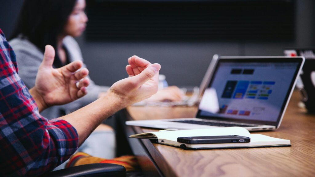 Person in plaid shirt gesturing during discussion with laptop and notebook on wooden desk