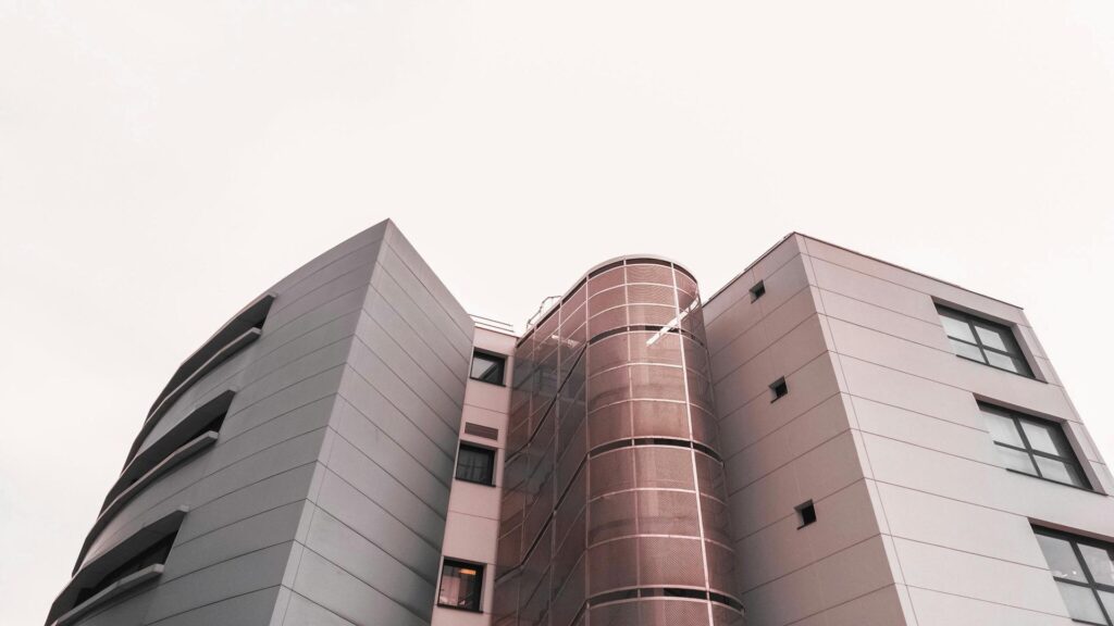 Low-angle view of a contemporary building featuring curved and angular facades in pink and gray tones with a cylindrical tower element