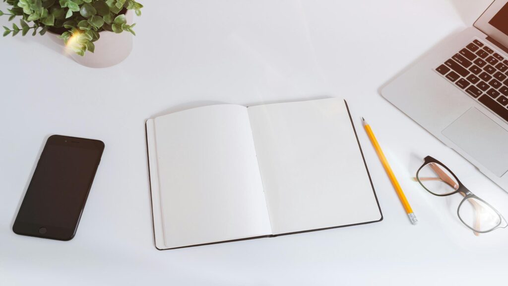 Minimalist desk setup with open blank notebook, smartphone, laptop corner, glasses and small plant