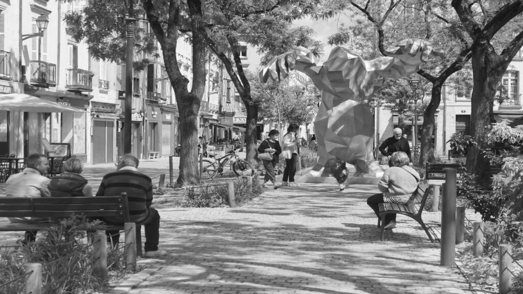 Black and white photograph of a modern geometric sculpture on a tree-lined street in Tours, France, with people sitting on benches and walking by