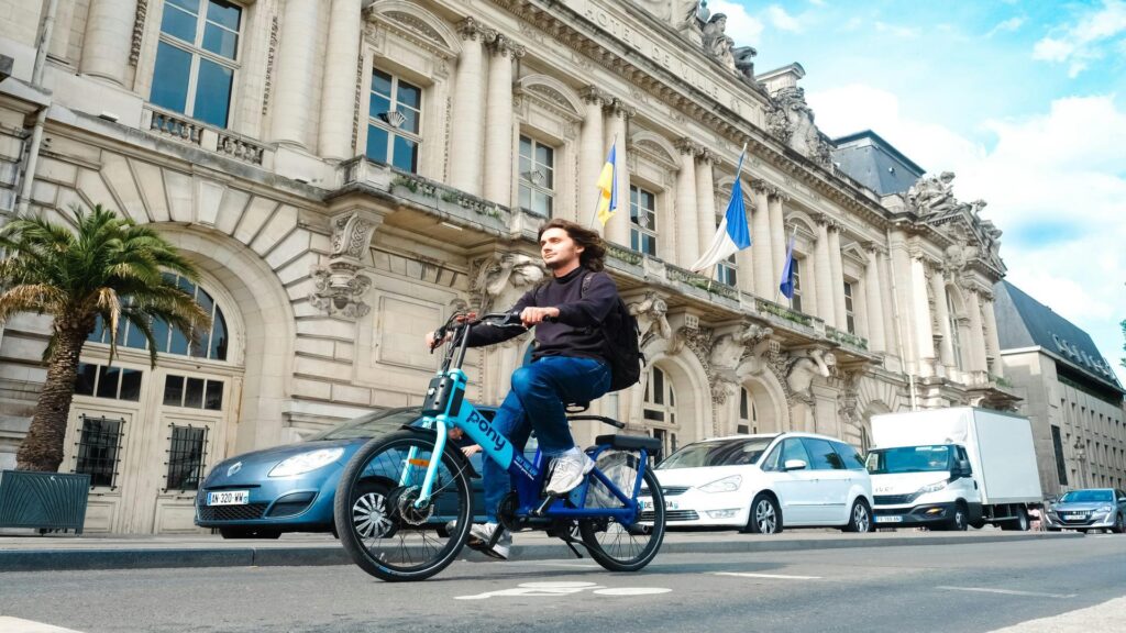 Person riding a blue Pony bike share bicycle in front of a grand Neoclassical building with palm trees and parked cars