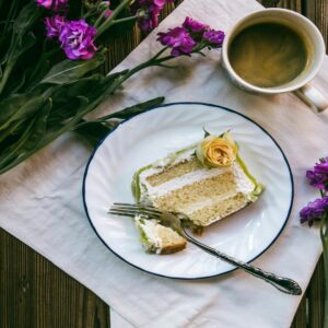 A slice of cake on a white plate with a cup of coffee and purple flowers on a wooden table.