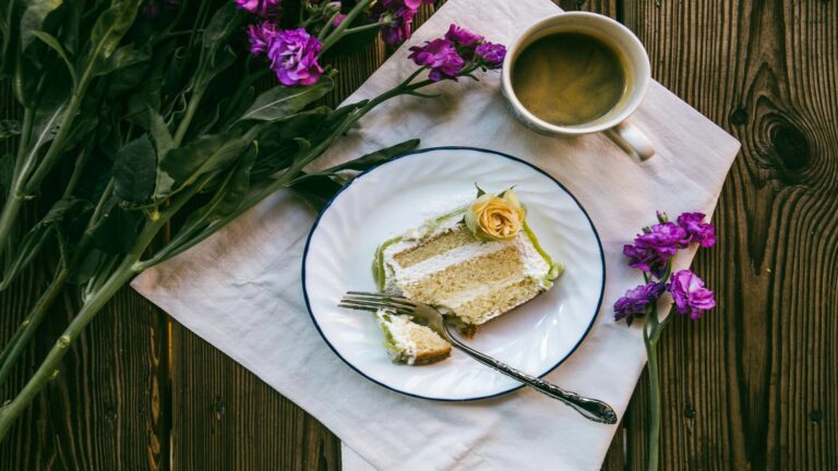 A slice of cake on a white plate with a cup of coffee and purple flowers on a wooden table.