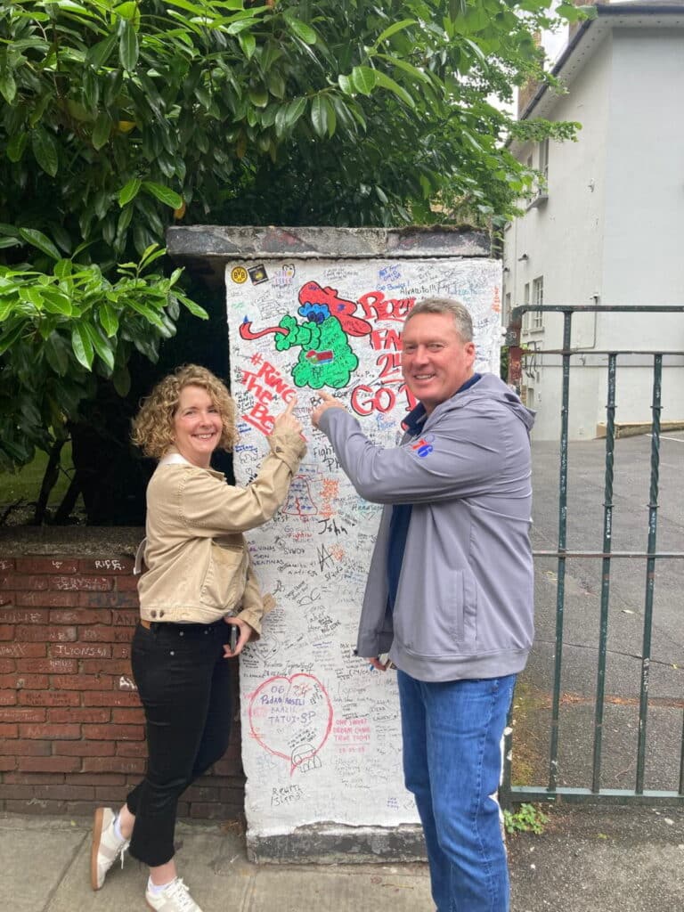 Two tourists enjoying a day trip from London, posing in front of a graffiti-covered wall at a famous sightseeing spot.