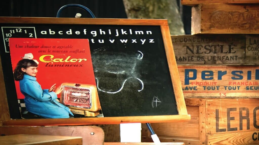 Nostalgic French classroom setting with chalkboard, vintage advertisements for Calor heater and Persil detergent, and wooden crates