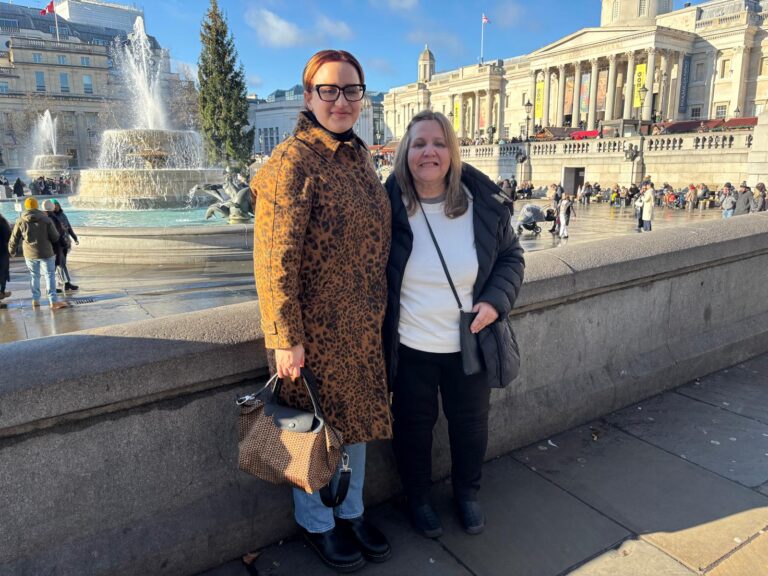 Two women posing in Trafalgar Square with fountains and historic buildings in the background, showcasing Great Places To Visit In London.