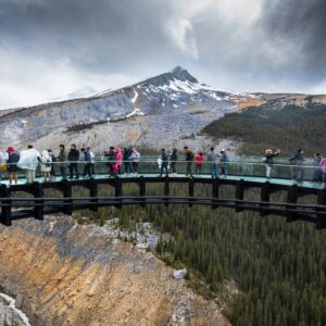 Tourists walking on a glass skywalk overlooking a canyon and mountain scenery.