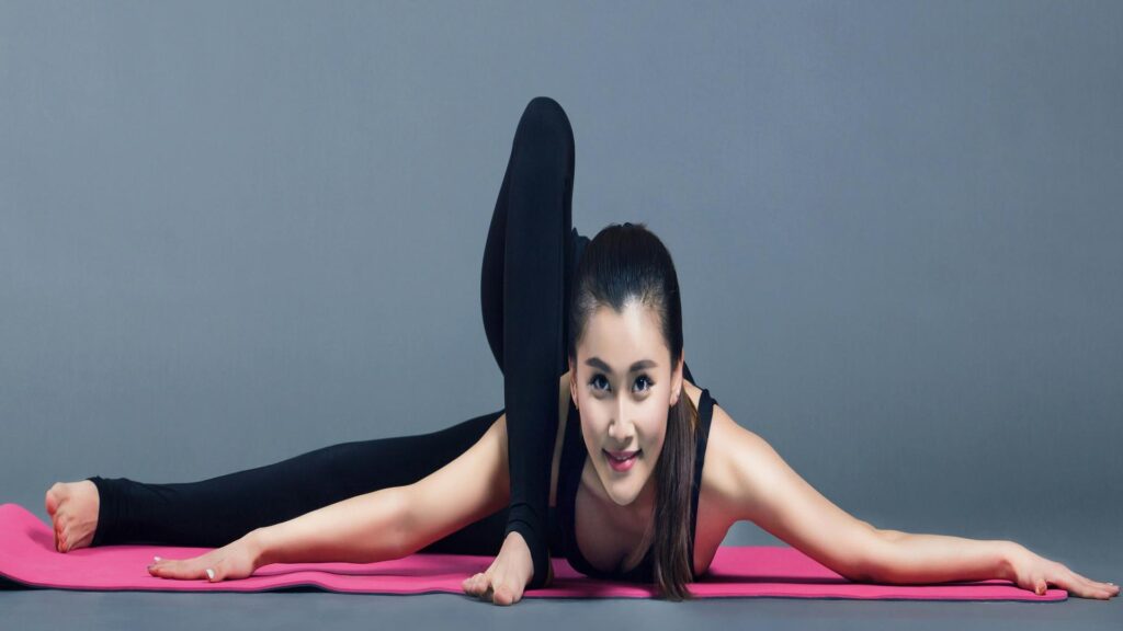 Woman in black athletic wear performing a split pose on pink yoga mat against grey background