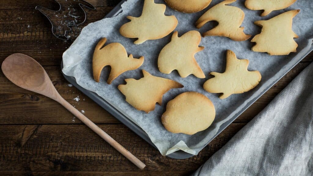 Freshly baked sugar cookies in various Halloween-themed shapes including cats, ghosts, and pumpkins on a baking sheet with parchment paper, wooden spoon on rustic wooden surface