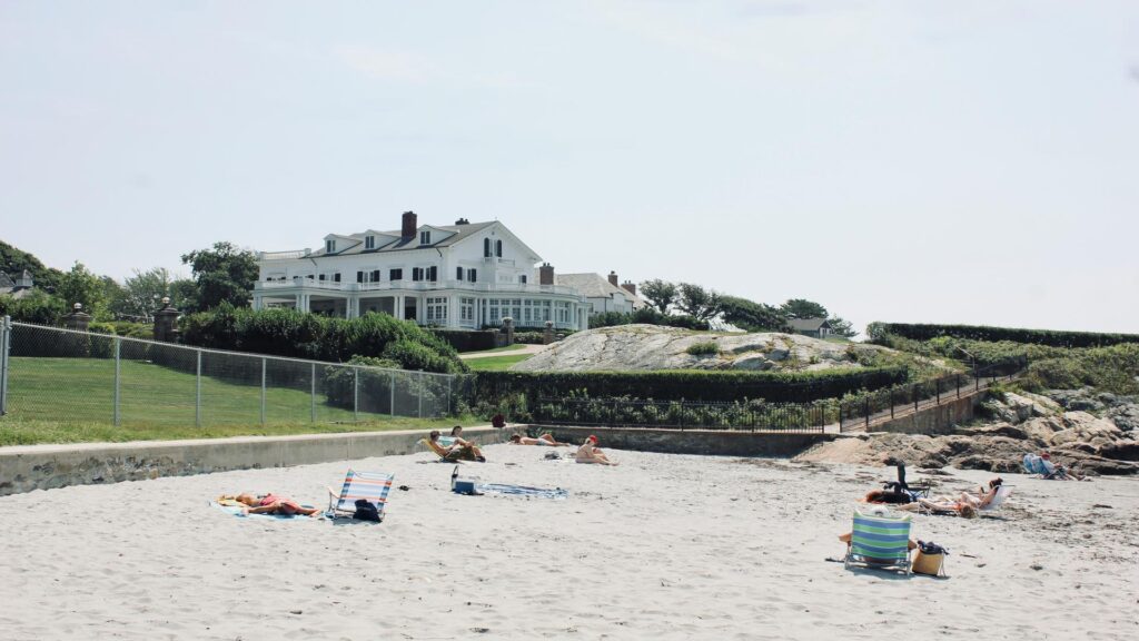 People sunbathing on a sandy beach with a large house in the background