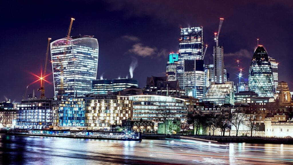 Night view of London's financial district showing illuminated skyscrapers including the Walkie-Talkie building and the Gherkin along the River Thames