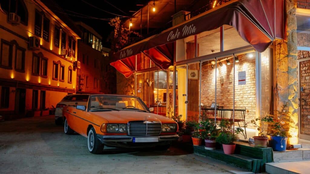 An orange vintage convertible car parked in front of a warmly lit cafe with a red canopy at night.