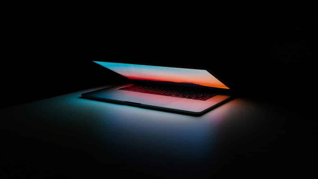 A partially closed laptop with a glowing screen displaying a colorful gradient in a dark room.