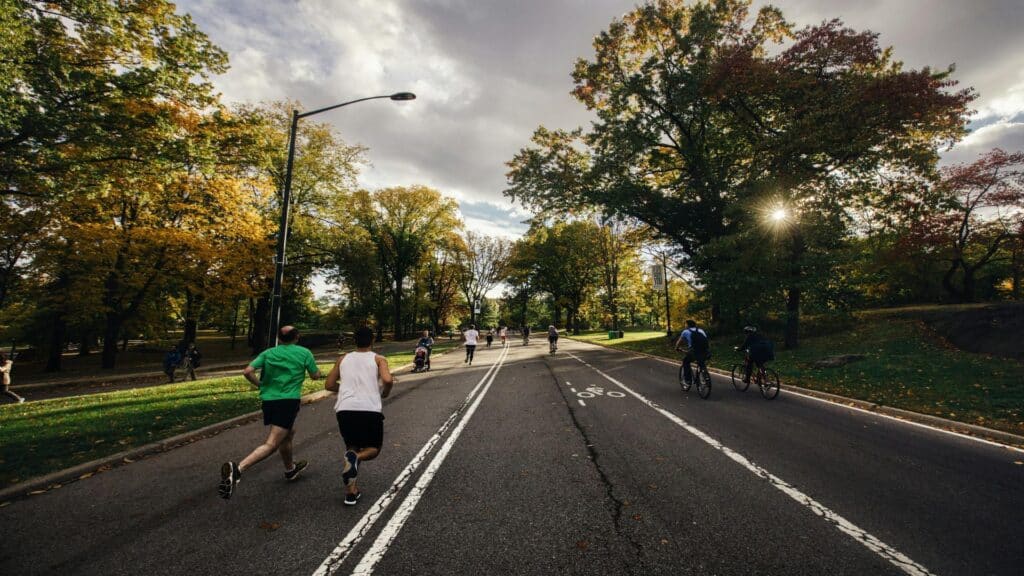 People jogging and cycling on a tree-lined road in Central Park during autumn, with golden foliage and dramatic cloudy sky