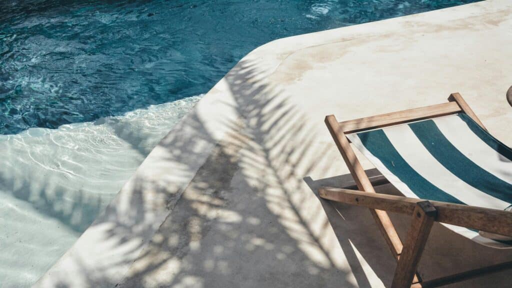 A striped deck chair by a pool with palm tree shadows.