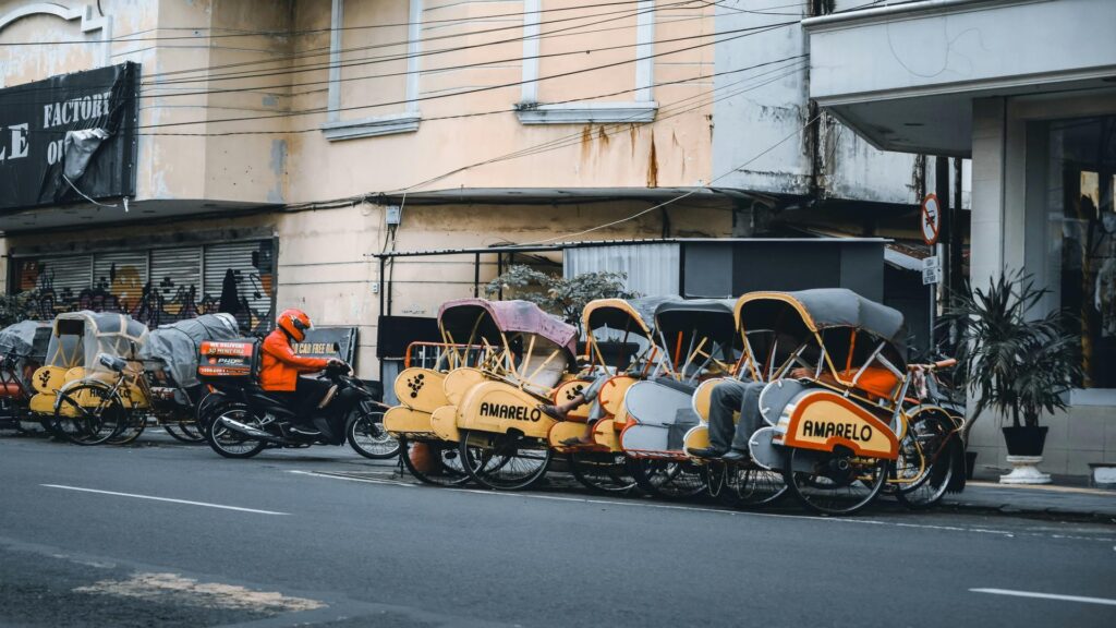 A row of colorful rickshaws parked on a city street with a delivery person on a motorcycle nearby.