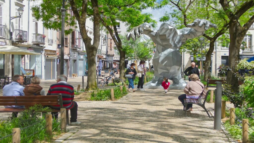 People sitting and walking near a large abstract sculpture in a tree-lined park