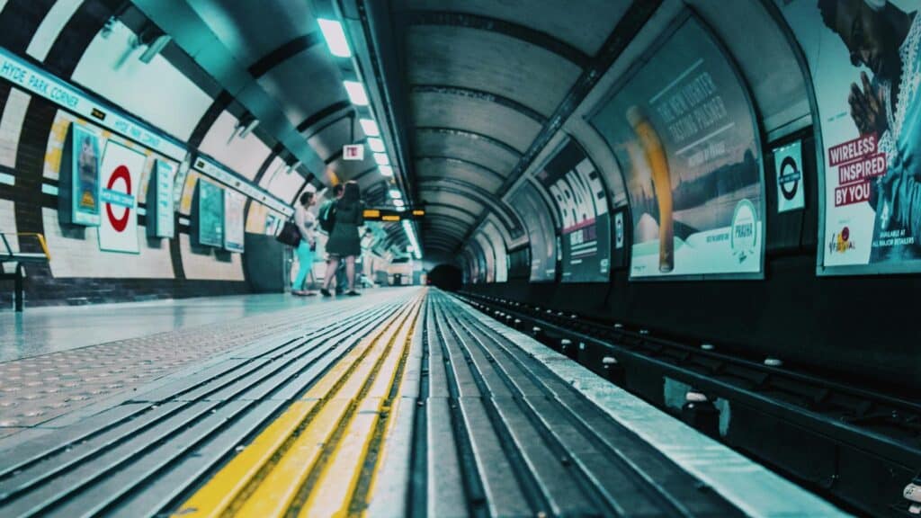 Low-angle view of a London Underground platform with tactile paving strips and advertising displays in a teal-tinted atmosphere