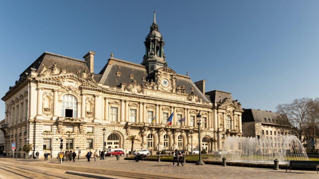 A grand historic building with a clock tower and fountain in front, captured on a sunny day.