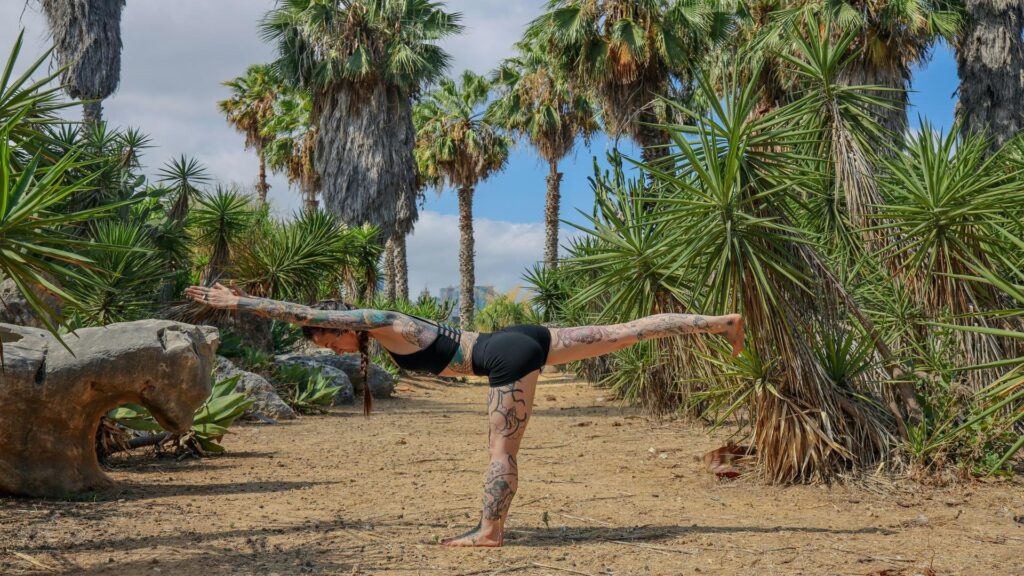 Person performing warrior III yoga pose in a tropical garden with palm trees