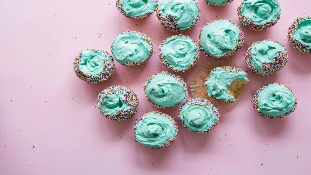 A collection of cupcakes topped with light blue frosting and rainbow sprinkles on a pink background.