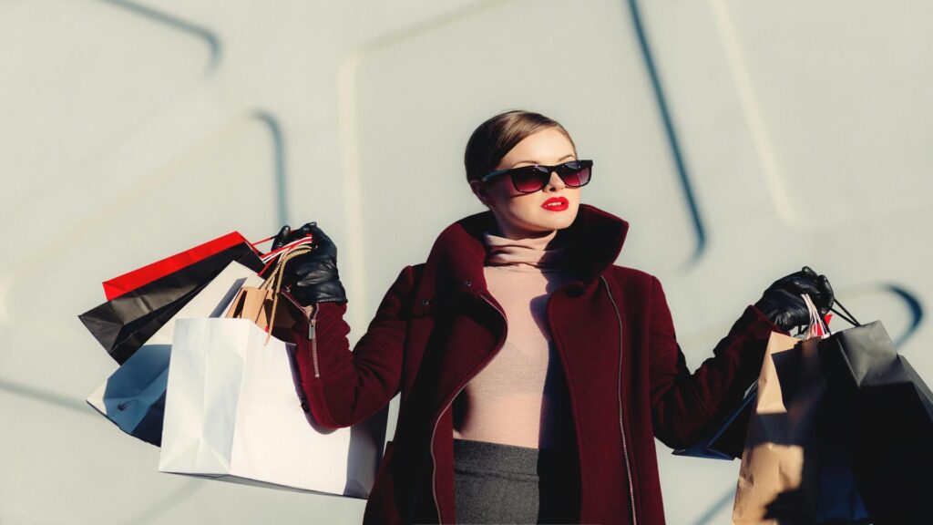 Fashionable woman in burgundy coat carrying multiple shopping bags against white wall