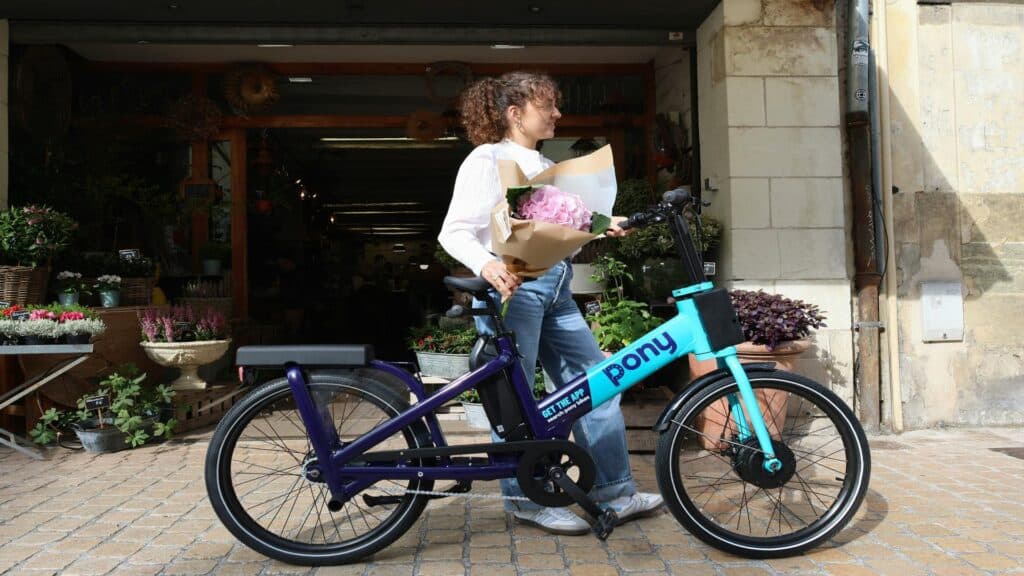 A woman holding flowers standing next to a Pony bike outside a flower shop.