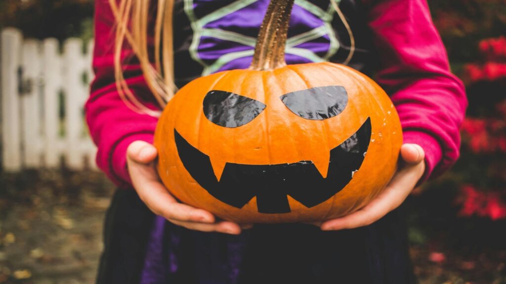 Person holding carved pumpkin with vampire-like face against purple and pink costume background