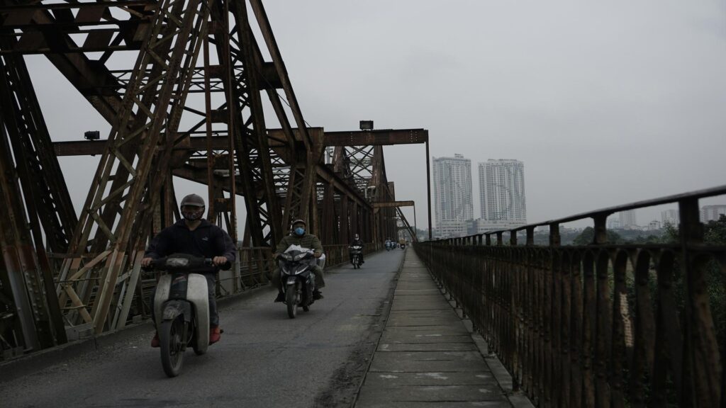 Motorcyclists riding across the Long Bien Bridge with high-rise buildings in the background.