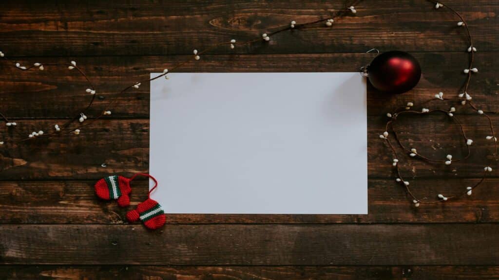 A blank white sheet surrounded by holiday decorations, including a red ornament, knitted mittens, and string lights on a wooden table.