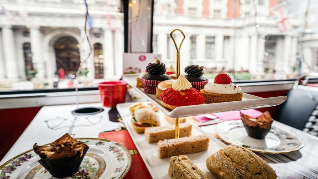 A tiered tray with assorted pastries, sandwiches, and scones on a table inside a bus with a city view outside.