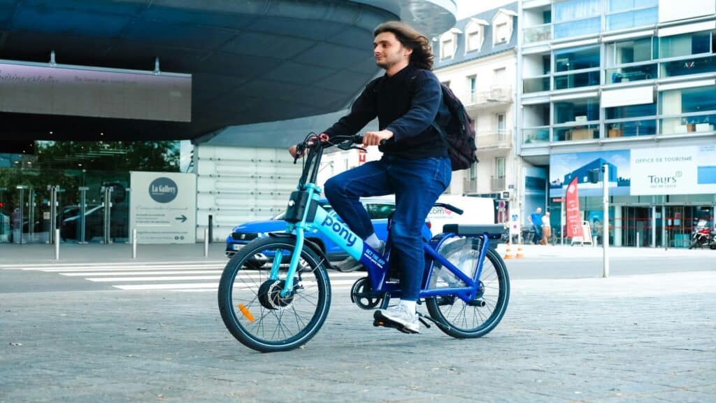 Person riding a blue Pony bike-sharing bicycle in front of Tours Tourist Office