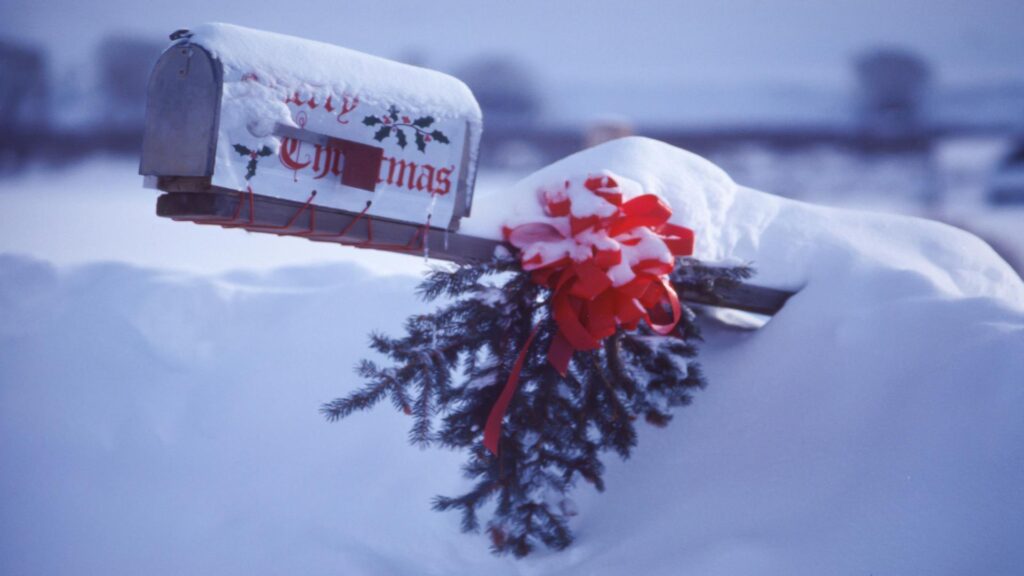 A mailbox decorated with Christmas-themed designs, red ribbons, and evergreen branches, covered in snow.