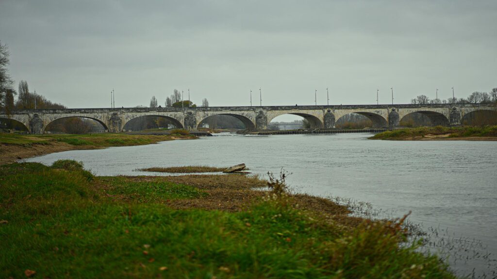 Stone arch bridge spanning across Loire River on cloudy day with grassy riverbank in foreground