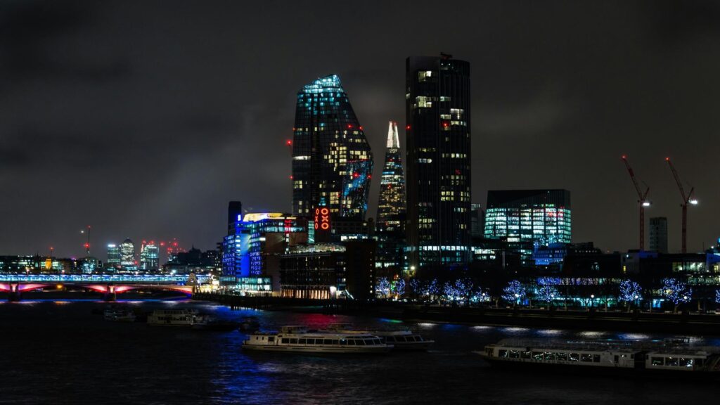 Nighttime cityscape of London showing illuminated skyscrapers along the Thames River with tourist boats