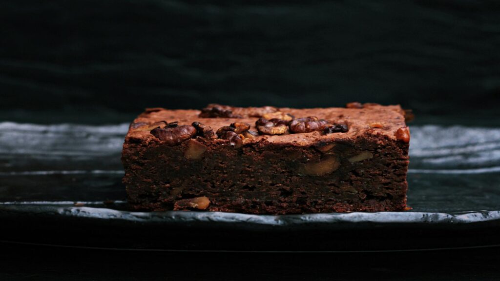 A rich chocolate brownie topped with nuts on a dark plate.