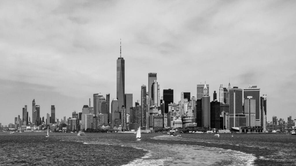 Black and white image of the New York City skyline with tall buildings and water in the foreground.