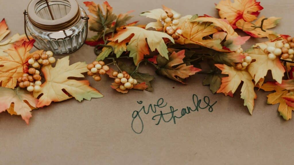 A decorative arrangement of autumn leaves, berries, and a glass jar with the words 'give thanks' written on a brown surface.