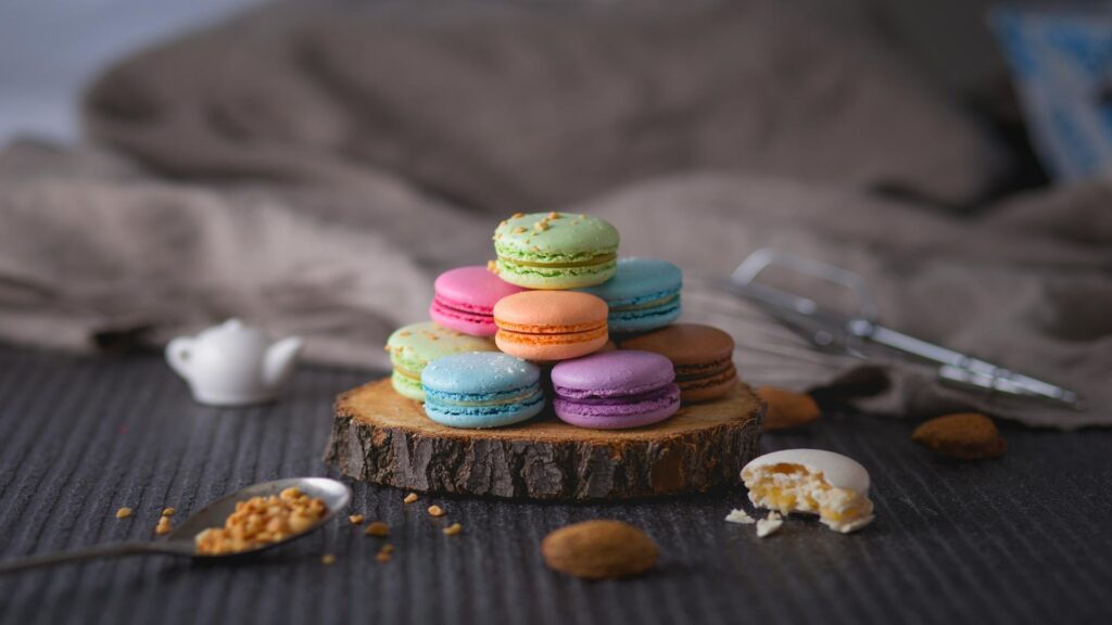 A stack of colorful macarons on a wooden base with scattered crumbs and a blurred background.