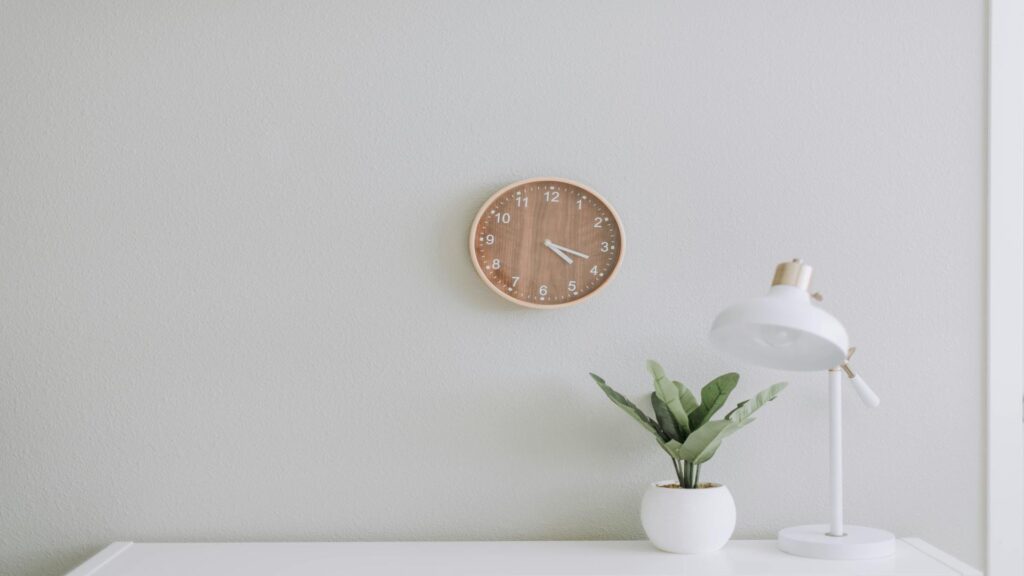 Minimalist workspace featuring a wooden clock, white desk lamp, and a green potted plant against a light gray wall.