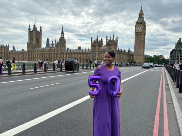 Woman holding purple balloons shaped as the number 50 on Westminster Bridge with Big Ben and the Houses of Parliament in the background, showcasing City Sightseeing London Reviews.
