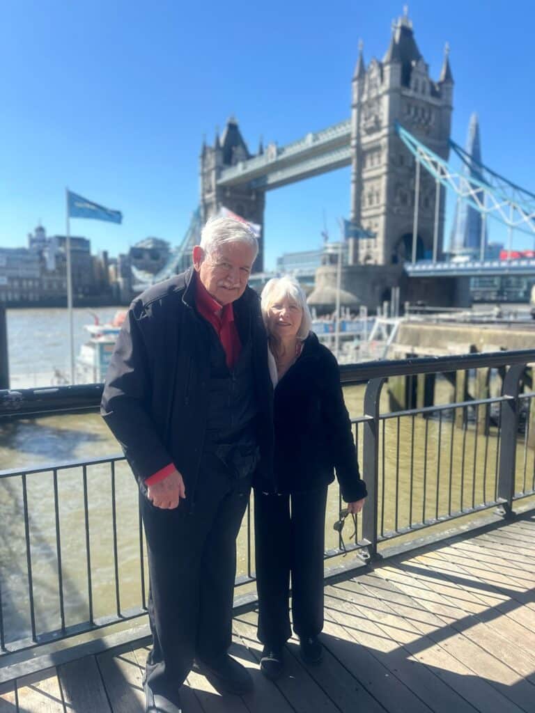 Elderly couple smiling during private walking tours London, standing in front of the iconic Tower Bridge with the Thames River backdrop for sightseeing